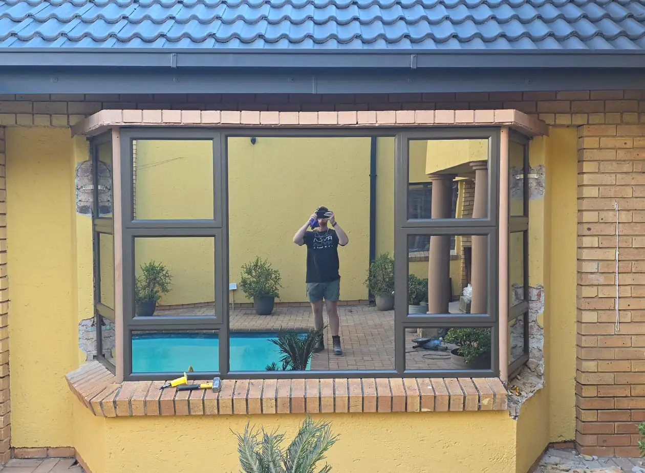 The same bay window rebuilt in charcoal aluminium with slim frames, clear glazing and no burglar bars, reflecting the swimming pool and courtyard behind the photographer.