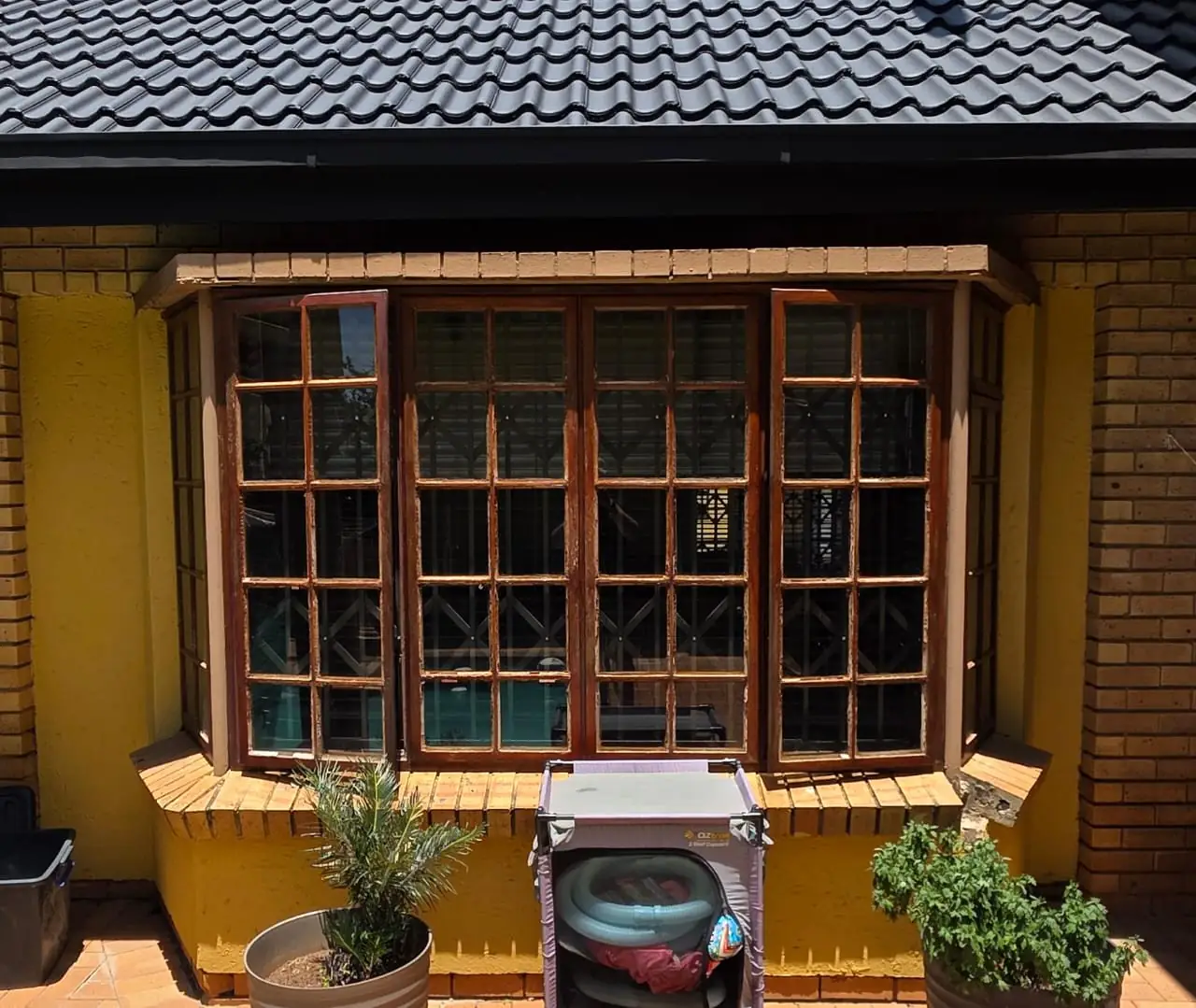 A bay window in dark varnished timber with peeling finish and diamond-pattern burglar bars, set into a yellow-painted wall above a face-brick sill.