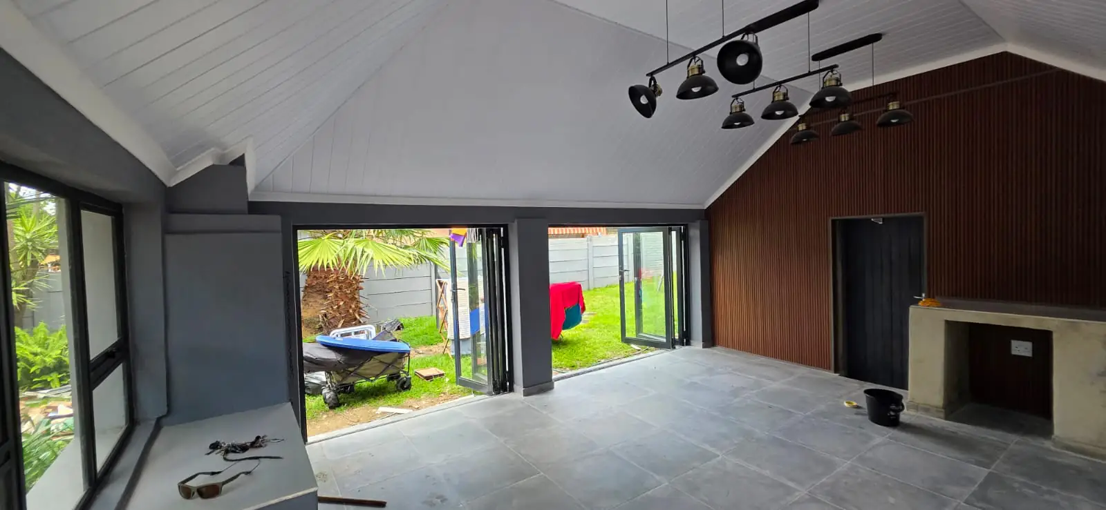 A patio converted into an enclosed entertainment room, with a pitched white ceiling, black track lighting and charcoal aluminium folding doors standing open onto a lawn.