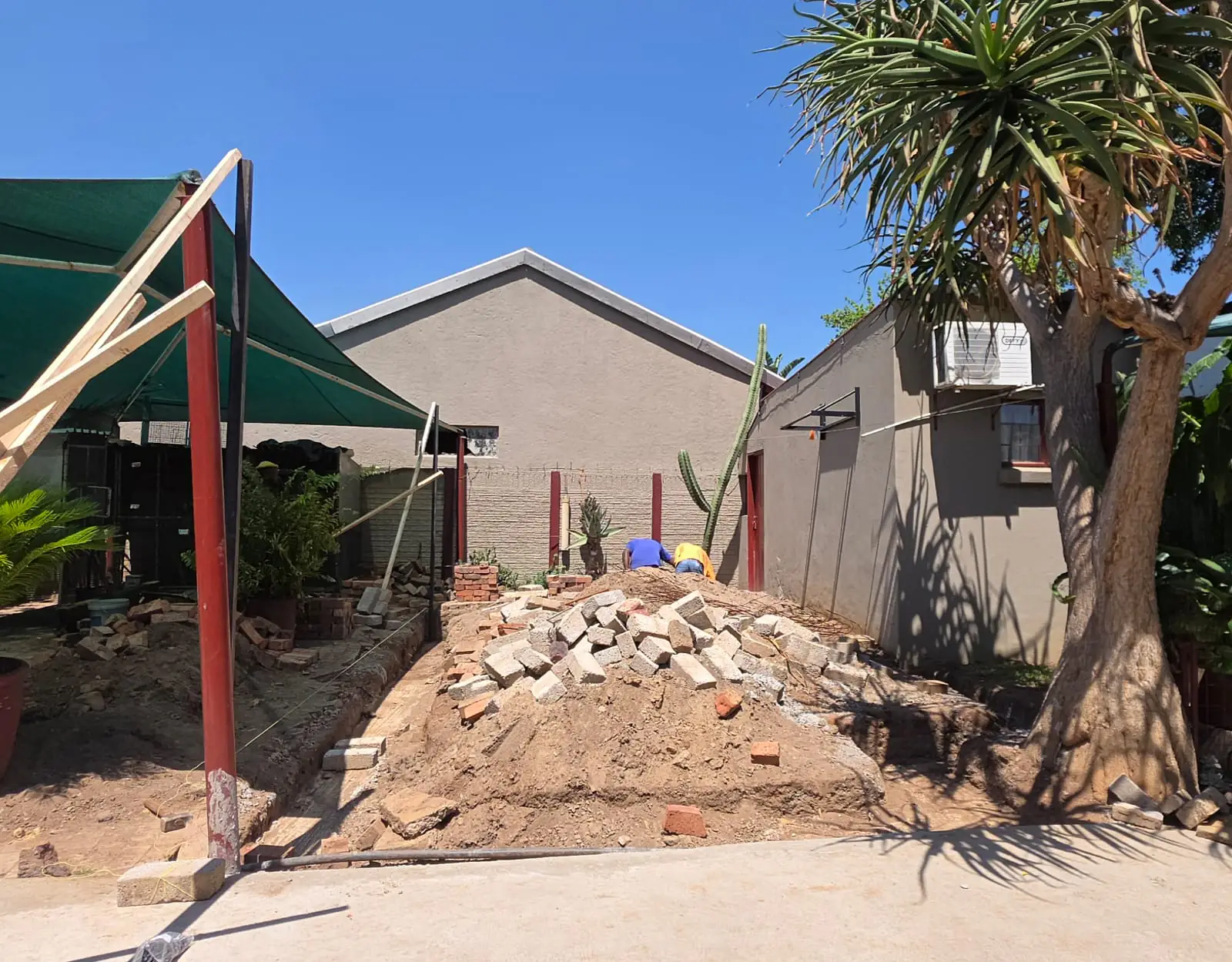 Excavated foundation trenches and heaps of sand and broken brick in a back yard, with red steel posts standing ready and a green shade sail overhead.
