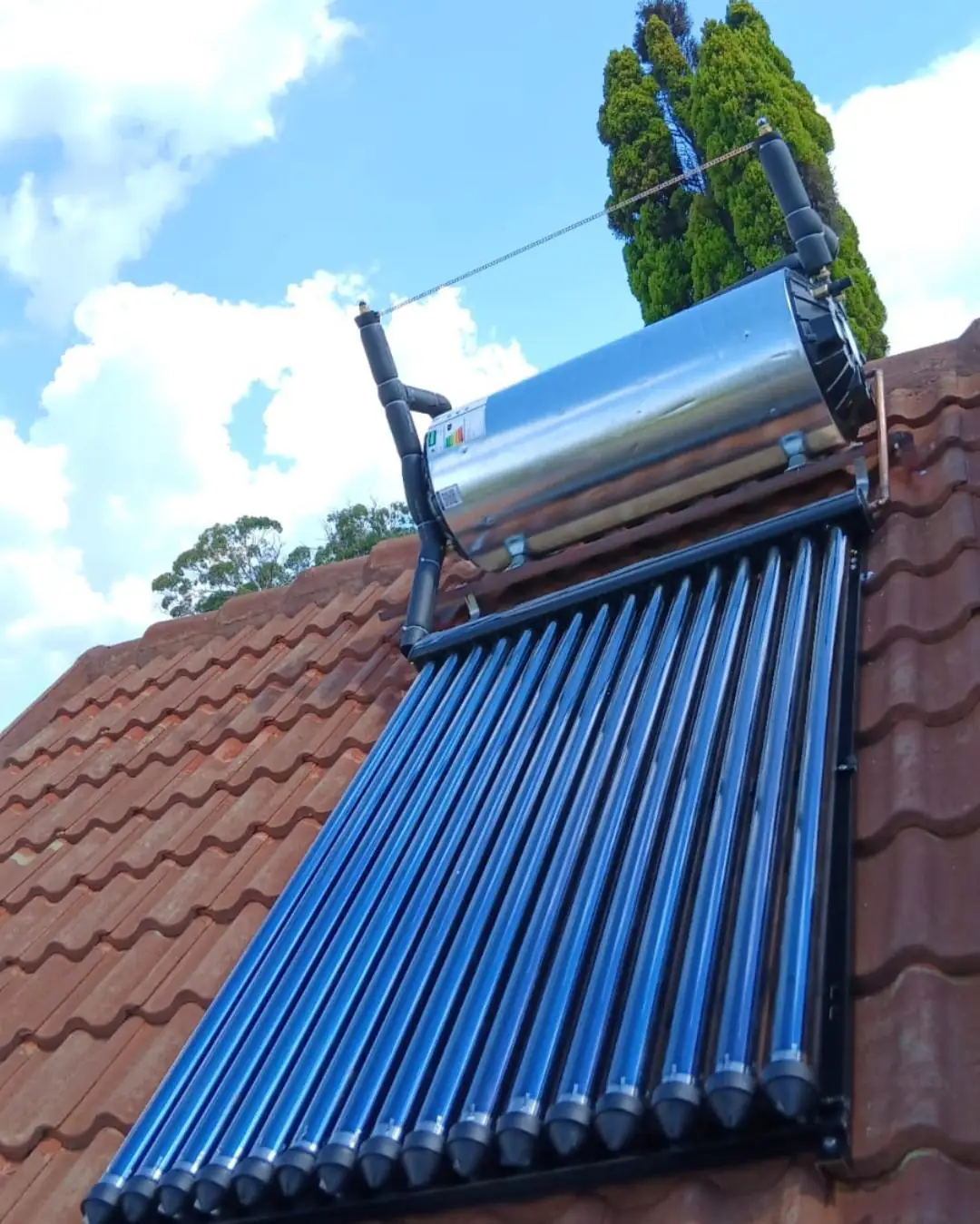 A stainless steel solar geyser and a bank of blue evacuated tube collectors mounted on a red tiled roof against a bright sky.